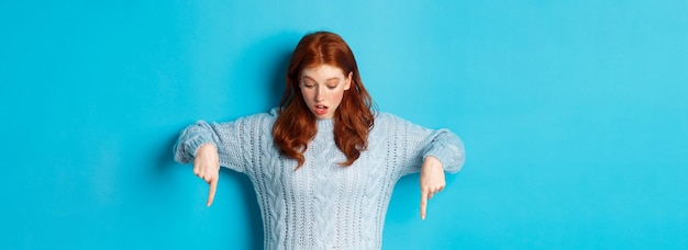 Free photo winter holidays and people concept impressed redhead girl in sweater looking and pointing down with