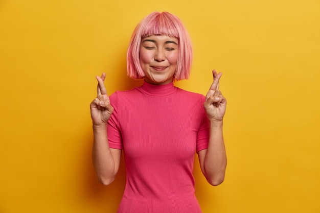 Wishful positive Asian woman crosses fingers before important event smiles happily stands with eyes shut has big hope in better