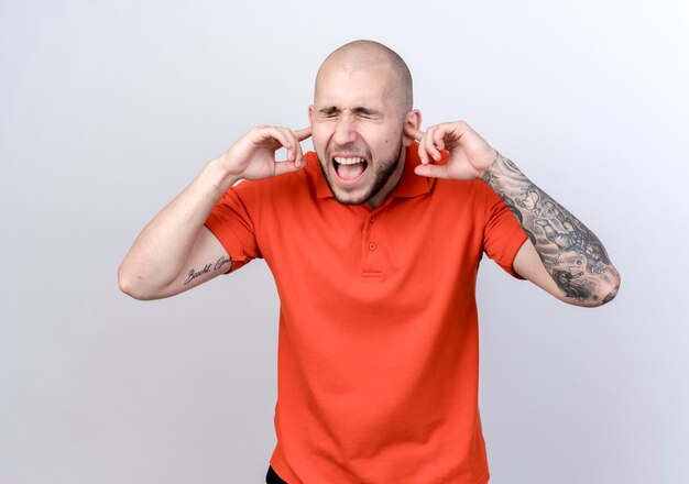 With closed eyes angry young sporty man closing ears isolated on white wall