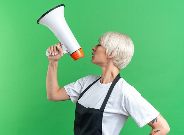 With closed eyes looking at side young beautiful female barber in uniform speaks on loudspeaker isolated on green background