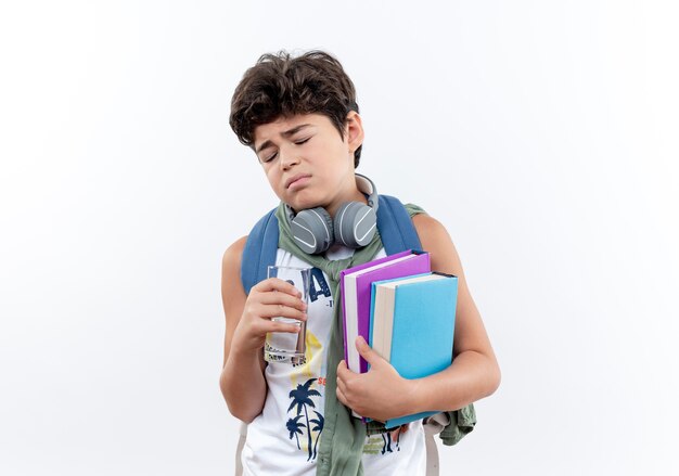 With closed eyes sad little schoolboy wearing back bag and headphones holding glass of water and book isolated on white