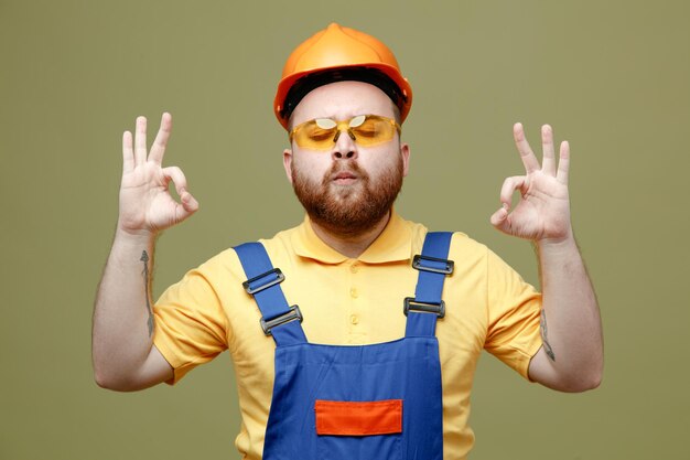 With closed eyes showing meditation young builder man in uniform isolated on green background