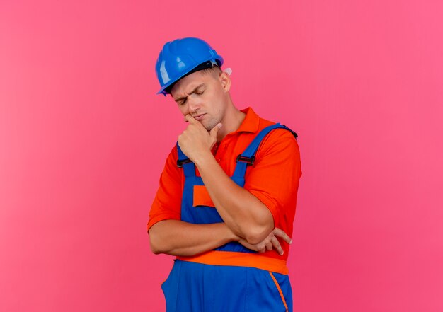 With closed eyes thinking young male builder wearing uniform and safety helmet putting hand under chin on pink