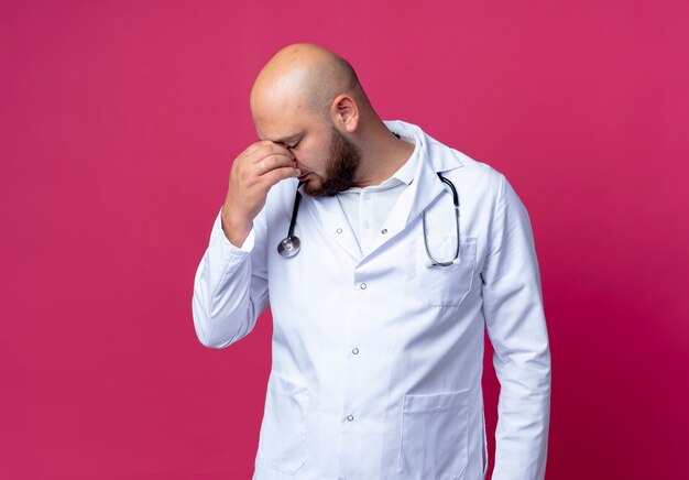 With closed eyes tired young bald male doctor wearing medical robe and stethoscope putting hand on nose isolated on pink with copy space