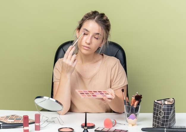 With closed eyes young beautiful girl sitting at desk with makeup tools applying eyeshadow with makeup brush isolated on olive green background