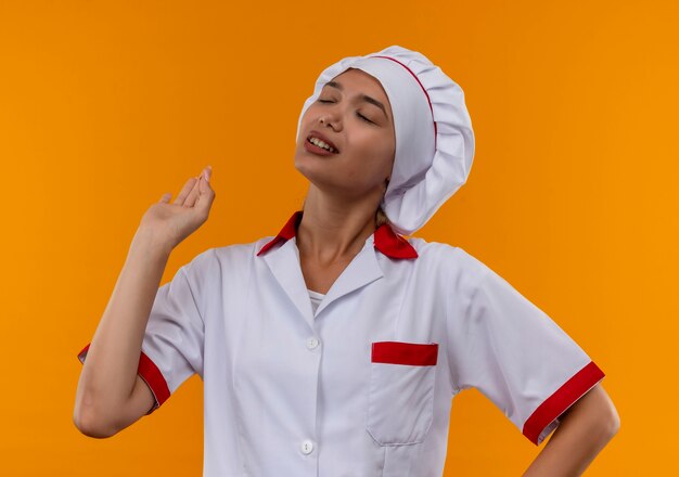 With closed eyes young cook female wearing chef uniform showing okey gesture on isolated orange background