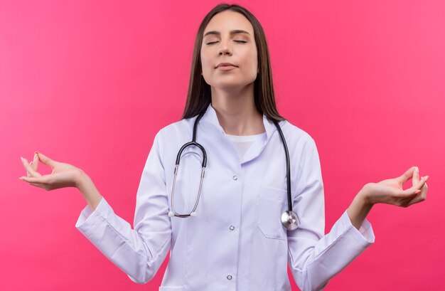 With closed eyes young doctor girl wearing stethoscope medical gown showing okey gesture on isolated pink wall