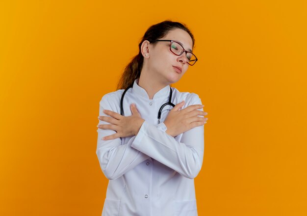 With closed eyes young female doctor wearing medical robe and stethoscope with glasses putting hands on shoulder isolated