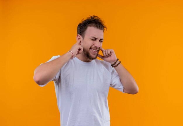 With closed eyes young guy wearing white t-shirt closed ears on isolated orange wall