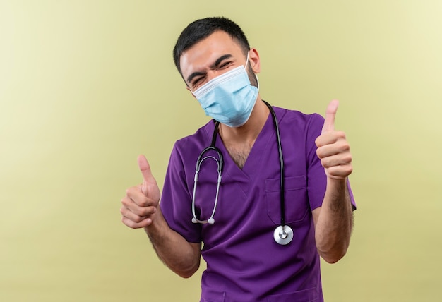 With closed eyes young male doctor wearing purple surgeon clothing and stethoscope medical mask his thumbs up on isolated green wall