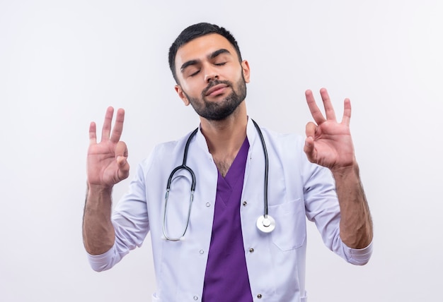 With closed eyes young male doctor wearing stethoscope medical gown showing okey gesture on isolated white