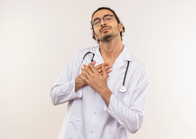 With closed eyes young male doctor with optical glasses wearing white robe with stethoscope putting hands on heart on isolated white wall with copy space