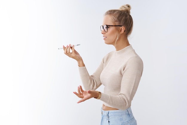 Woman arguing on phone talking via speaker holding smartphone near mouth spread hands in dismay bothered and intense having serious argument sending voice message on white