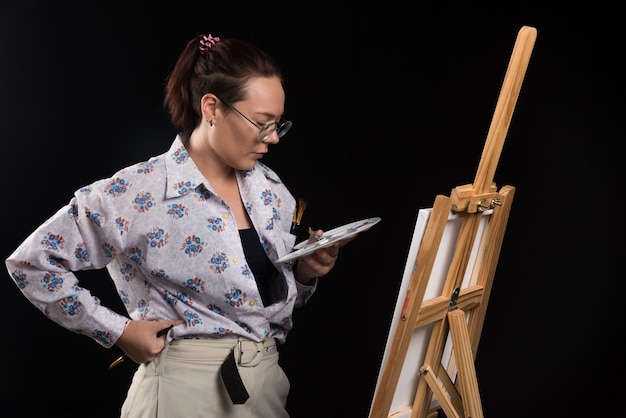 Woman artist holding brush and looking at canvas on black background