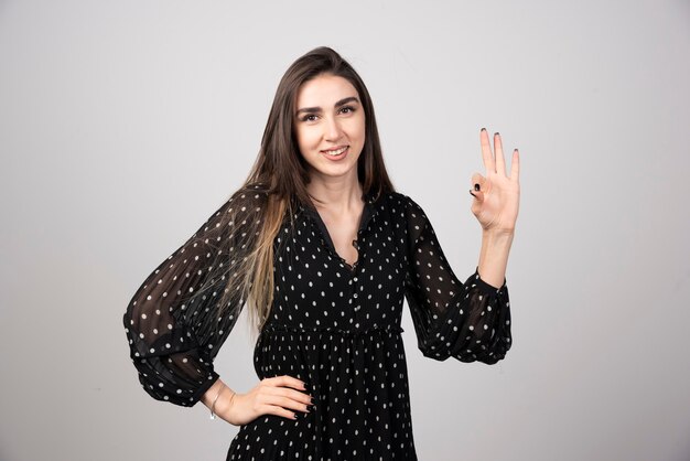 Woman in black dress showing ok sign on gray.