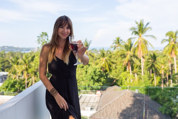 Woman in black evening dress with glass of wine on tropical balcony