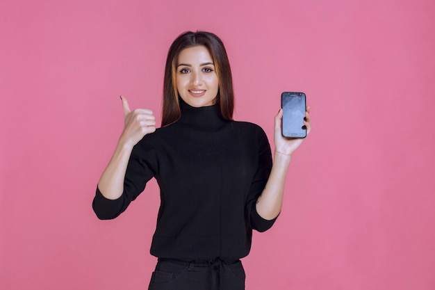 Woman in black sweater holding a smartphone and making thumb up. 