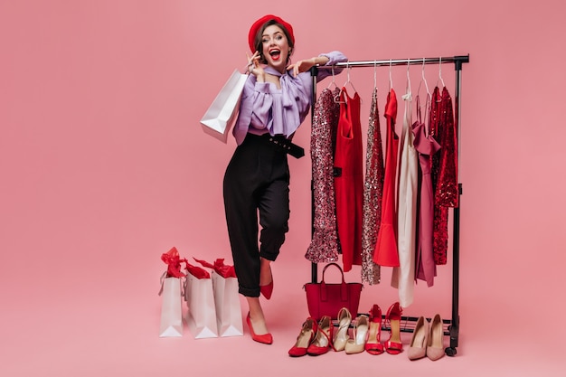 Free Photo woman in black trousers and purple blouse laughs, leaning on stand with elegant clothes on pink background.