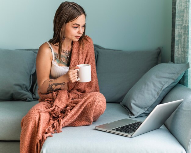 Woman in blanket having coffee and working on laptop at home during the pandemic