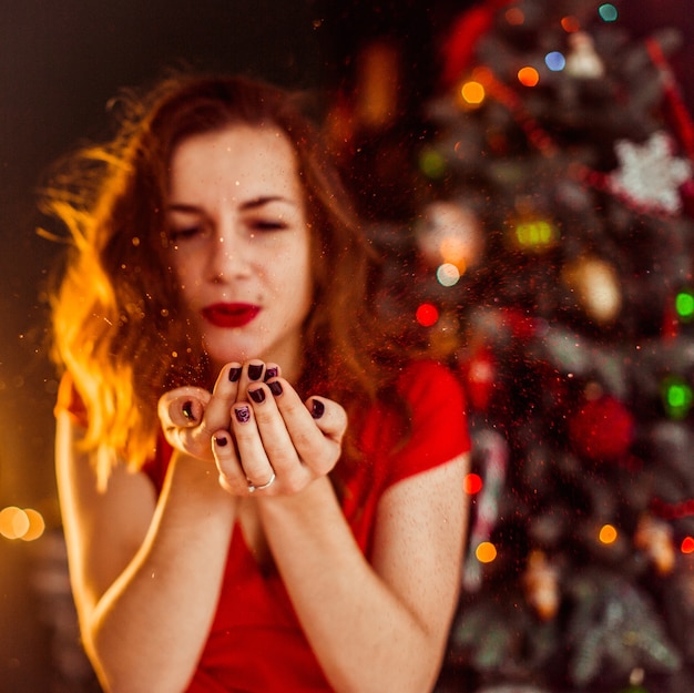 Woman blows snow from her palms standing before the Christmas tree 