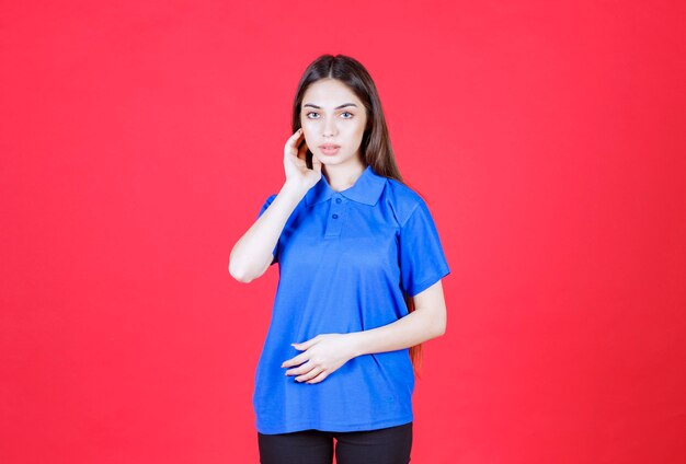 woman in blue shirt standing on red wall and looks confused and thoughtful. 