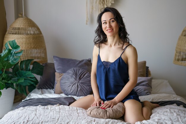 A woman in a blue slip sits on the bed and smiles.