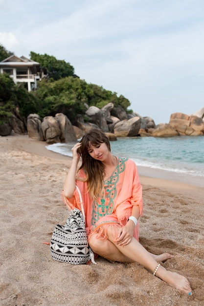 Woman in boho summer dress sitting on sand near sea. Tropical mood.