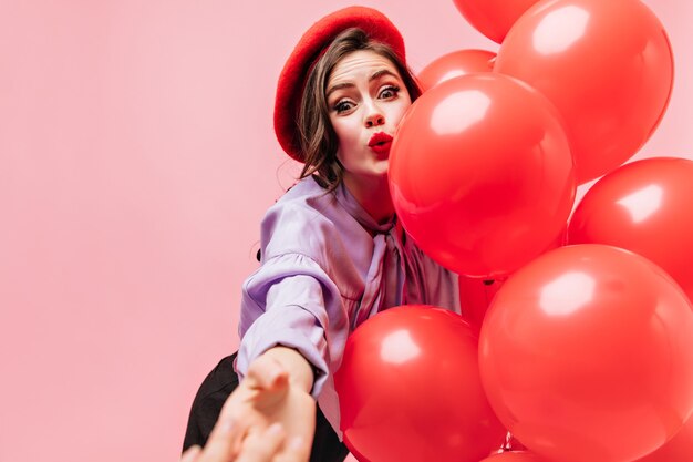 Free photo woman in bright beret and purple blouse is looking at in surprise, reaching for camera and holding balloons.