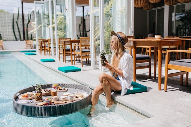 Free photo woman in brown hat drinking pineapple cocktail while resting near pool. magnificent blonde female model chilling at resort cafe in weekend morning.