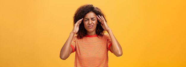 Woman cannot recall important information looking intense trying concetrate under pressure feeling tensed touching temples closing eyes while thinking hard posing against orange background