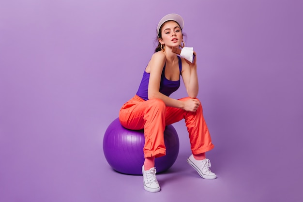 Free photo woman in cap and sporty purple-orange suit sits on fitball and holds milk chocolate