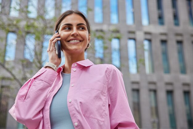  woman in casual clothes uses smartphone for conversation stands at street near modern building looks away happily walks in urban setting enjoys leisure time with gadget