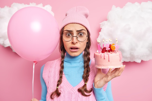 Free photo woman celebrates her 26th birthday holds cake with burning candles and inflated balloon wears pink hat turtleneck big round spectacles stands indoor