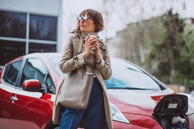 Woman charging electro car at the electric gas station and drinking coffee