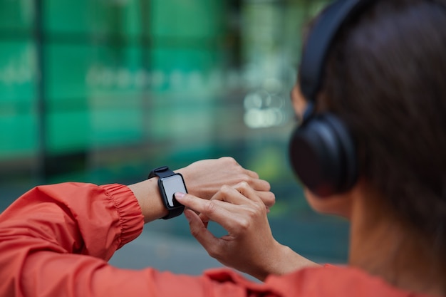 Free photo woman checks results of fitness training on smartwatch listens music via headphones dressed in anorak poses on blurred