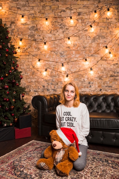 Free photo woman next to christmas tree with teddy bear wearing christmas hat