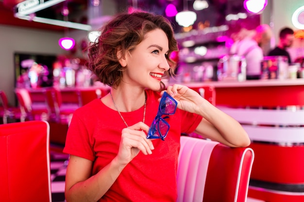 woman in colorful outfit in retro vintage 50's cafe sitting at table wearing red shirt, blue sunglasses having fun in cheerful mood, red lipstick make-up