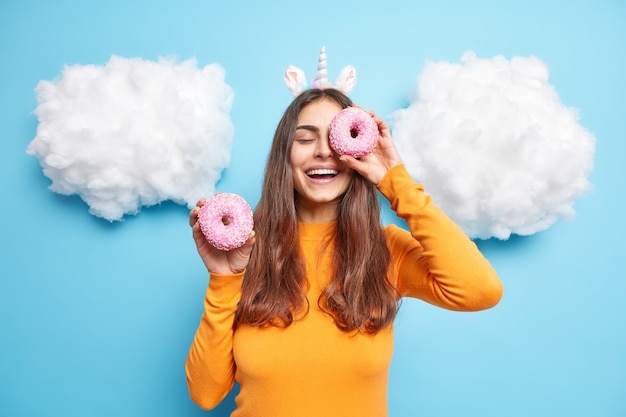 Free photo woman cons eyes with glazed delicious donuts smiles positively has sweet tooth wears orange jumper isolated on blue