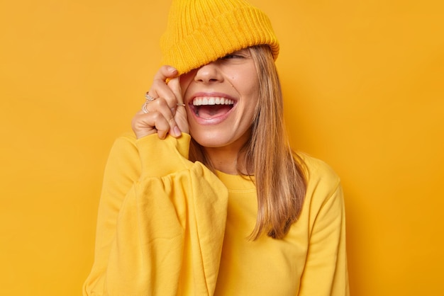 Free photo woman cons eyes with hat has joyful expression dressed in casual jumper isolated on bright yellow. carefree teenage girl foolishes around has funny expression