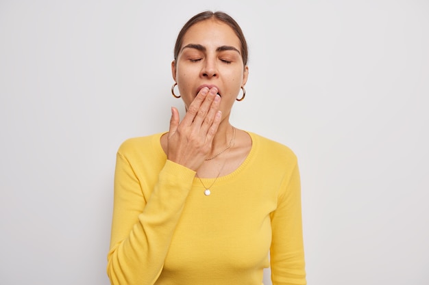 Free Photo woman cons mouth with hand keeps eyes closed wears casual yellow jumper feels fatigue or sleepy stands on white