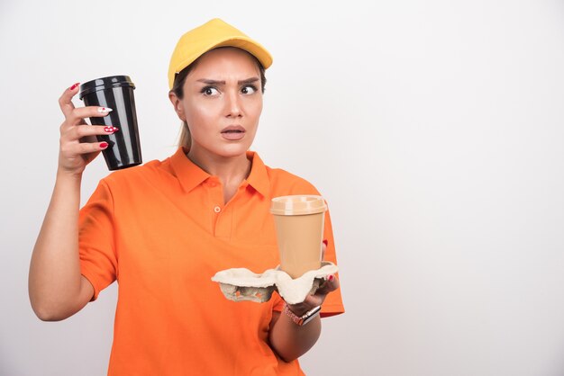 Woman courier holding two cups of coffees on white wall. 