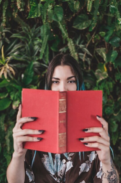 Woman covering her mouth with book standing against growing plant