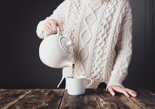 Free photo woman in cozy warm white thick knitted sweater pours hot tea from big ceramic teapot to blank cup