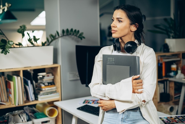 Woman designer with musical earphones standing in office holding folder