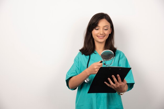 Woman doctor looking at clipboard with magnifying glass on white background.