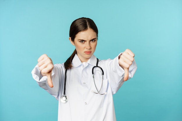 Woman doctor medical worker showing thumbs down and frowning angry dislike disappove smth standing i...