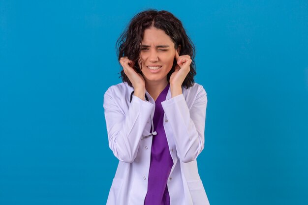 woman doctor wearing white coat with stethoscope coning ears with fingers with annoyed expression for the noise of loud sounds on isolated blue