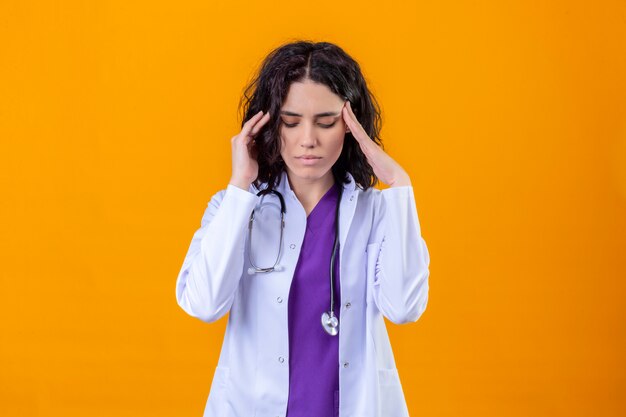 woman doctor wearing white coat with stethoscope looking unwell touching temples having strong headache standing on isolated orange