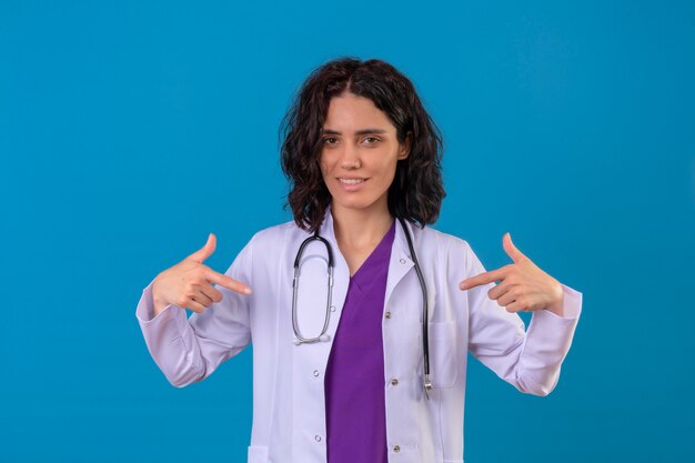 woman doctor wearing white coat with stethoscope pointing at herself with thumbs looking confident on isolated blue