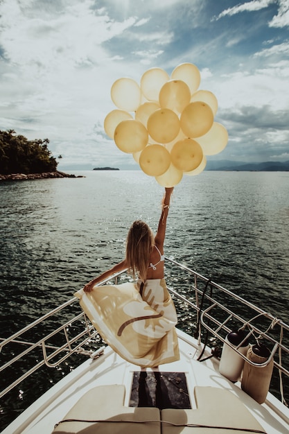 Free photo woman in a dress standing on a yacht, holding golden balloons while sailing